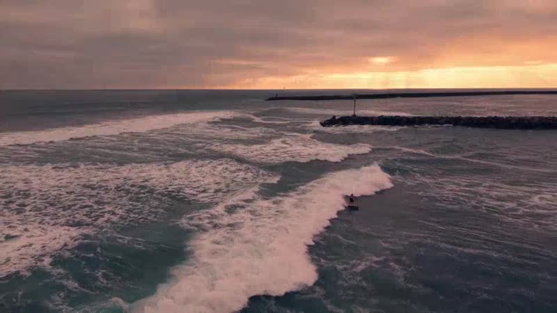Aerial footage of a surfer riding a wave beside the jetty at dusk
